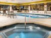 Indoor pool area with chairs and tables surrounding a larger pool and a smaller hot tub. Large windows line the walls.