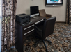 Office setup with a dark desk, computer, printer, and office chair on a patterned carpet. Brown armchair and wall-mounted screen visible in the background.
