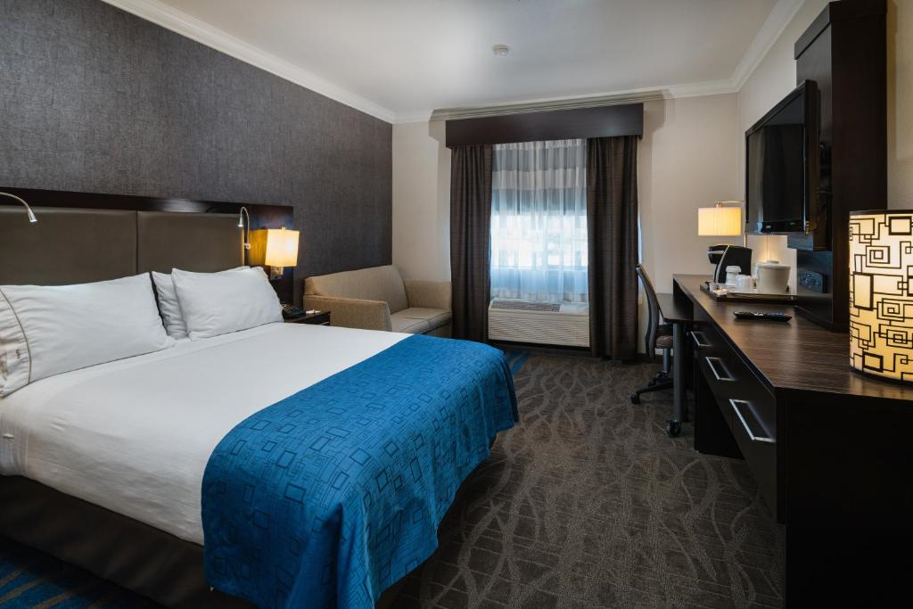 Hotel room with a large bed, blue bedspread, and a sofa. The room includes a desk with a TV, lamp, and coffee maker, and a window with curtains.