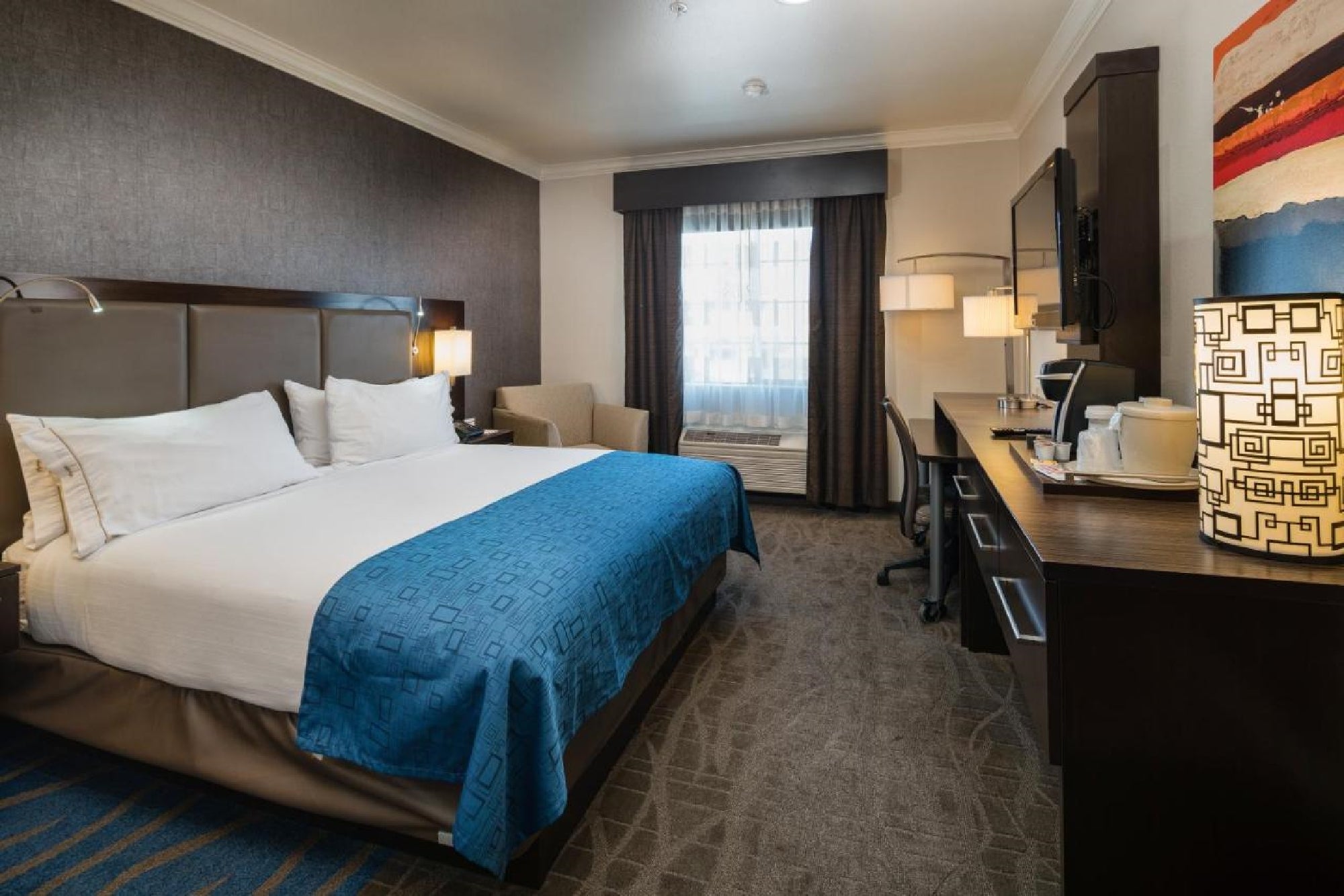 Hotel room with a king-size bed, blue blanket, desk, chair, and TV. A window with curtains and a lamp are visible.