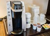 Single-serve Keurig coffee maker with K-cups, disposable cups, lids, and condiments on a countertop.