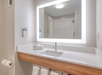 Bathroom with a white countertop, under-mount sink, illuminated mirror, towel rack with towels, soap dispensers, and a shower visible in the background.