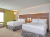 Hotel room with two double beds, white bedding, wooden headboard, and a lamp on the wall. A window with closed blinds is on the left. Neutral-colored carpet and walls create a simple decor.