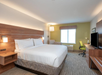 A modern hotel room with a large bed, wooden headboard, desk with chair, armchair, TV, and a window with natural light.