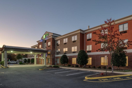 Exterior view of a three-story hotel with a covered entrance, surrounded by trees. The parking area is partially visible, with no vehicles parked near the entrance.