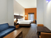 Hotel room with a large bed, blue sofa, wooden tables, and chairs. The room has an orange accent wall, a large window, and a workspace.