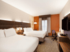 Hotel room with two double beds, a desk with a chair, a wall-mounted TV, and an orange accent wall. Large window with curtains on the right.