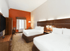 Hotel room with two double beds, a TV, desk, and chair. Room features an orange accent wall, window with curtains, and modern lighting.