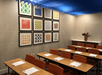 Classroom-style meeting room with rows of tables and chairs, note pads on each table, colorful geometric art on the wall, and a flower vase on a sideboard under a blue ceiling feature.