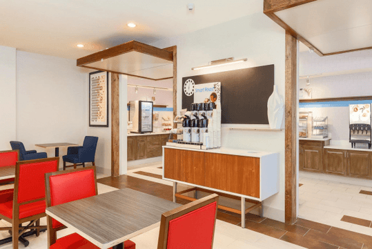 A hotel breakfast buffet and dining room with wooden tables with red chairs, a coffee bar, and a full buffet in the background.