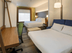 A hotel room with two white beds, a deck and office chair, a green accent wall, several lamps, and a curtained window on the far wall.