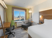 A hotel room with a white bed, a desk and office chair, a lounge chair, a lamp and side table, and window showing the city of Las Vegas.