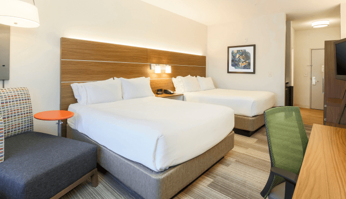 A modern hotel room with two white beds, a night stand with two lamps, an accent chair, a desk and office chair, and a painting on the wall.