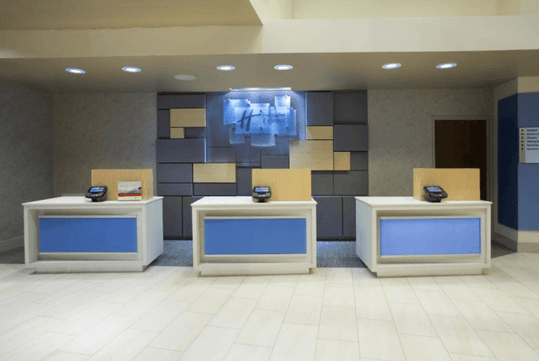 Hotel reception area with three desks, each equipped with a card reader. A sign displaying "Holiday Inn Express" is mounted on the patterned wall behind the desks.