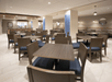 Empty dining area with wooden tables and chairs, beige tile flooring, and neutral-colored walls. Round lamps hang from the ceiling, and booths line the walls.