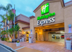 Holiday Inn Express entrance with a warmly lit exterior, American flag, palm trees, and a parking area with cars.
