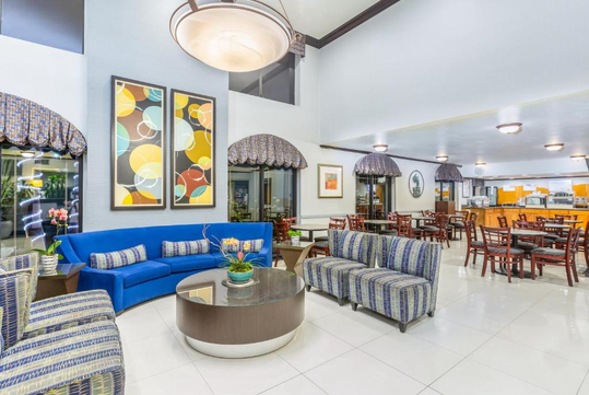 Modern hotel lobby with patterned sofas, a blue couch, and a glass coffee table. Colorful abstract art on the walls. Dining area with wooden tables and chairs in the background.