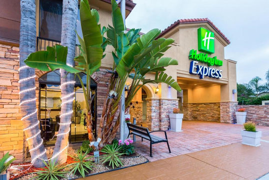 Exterior view of a Holiday Inn Express hotel entrance with a lit sign, tropical plants, and potted greenery along a stone pathway.