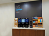 Coffee station with two coffee dispensers, a hot water dispenser, and assorted creamers and cups on a wooden counter against a black wall with the text "Must. Have. Coffee. EX-press.