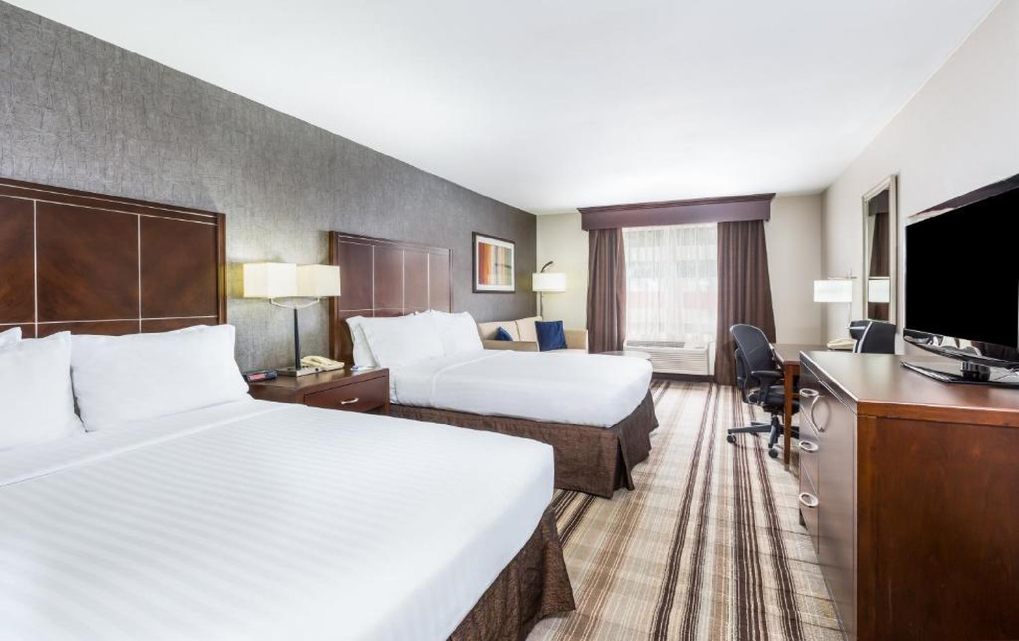 Hotel room with two double beds, bedside tables with lamps, a flat-screen TV, desk with chair, and a window with curtains. The decor features neutral tones and modern furnishings.