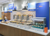 Buffet breakfast area with chafing dishes labeled for hot food and trays of pastries on a counter. Blue signs above display breakfast options like fruit and cereal.