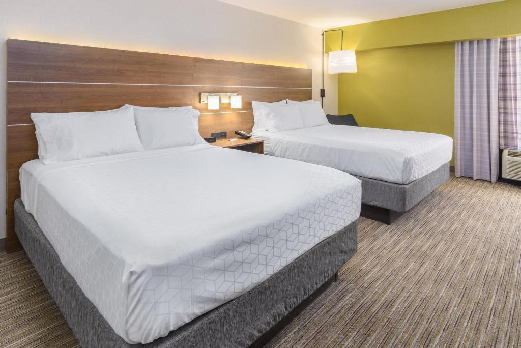 Hotel room with two double beds, a wooden headboard, white bedding, a nightstand with lamps, a green accent wall, and striped carpet flooring. Curtains cover the window on the right.