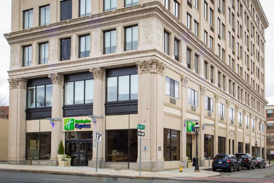 A multi-story building houses a Holiday Inn Express on a street corner. Cars are parked along the side of the street.
