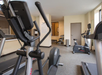 A gym room with exercise equipment, including a stationary bike, elliptical machines, and a treadmill, on a carpeted floor with beige walls and natural light coming through the windows.