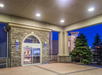 Entrance of a building with stone façade, automatic doors, and flanked by evergreen trees under a covered area at dusk.