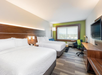 Modern hotel room with two double beds, white bedding, a large window, desk, chair, and wall-mounted TV. Decor features wood accents and green highlights.