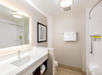 Modern bathroom with a white sink, illuminated mirror, abstract wall art, and towels. Shower area with toiletries in the corner. Neutral color scheme.