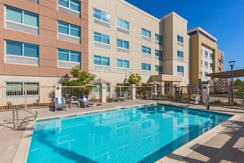 Outdoor swimming pool in front of a modern multi-story hotel building with beige and brown exterior. The pool area includes lounge chairs and landscaping.