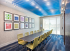 A conference room with a long table, yellow chairs, and colorful abstract artwork on the wall. Blue ceiling accents and large windows let in natural light.