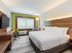 A modern hotel room with a large bed, wood furnishings, flat-screen TV, desk with chair, and a window with a green accent wall.