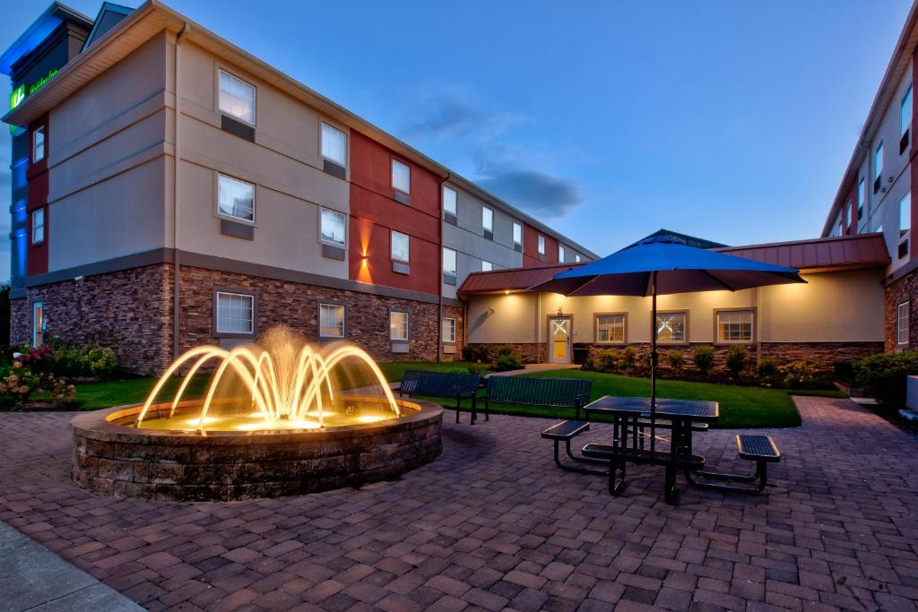 Courtyard with a lit fountain, picnic tables, benches, and a large blue umbrella, surrounded by buildings at dusk.