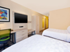 Hotel room with two double beds, a wall-mounted TV, a desk with a green chair, and a wardrobe. The room has modern decor with a colorful abstract painting.