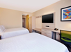 Hotel room with two double beds, a wall-mounted TV, a desk with a chair, and a colorful abstract artwork on the wall. The room has light-colored walls and furnishings.