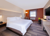 A hotel room with a king-size bed, nightstands, a desk with a chair, a window, and an armchair. The room features neutral tones with an orange accent wall.