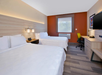 Hotel room with two double beds, white bedding, a desk with a green chair, a flat-screen TV on the wall, and a window with a view. Warm lighting and an orange accent wall enhance the decor.