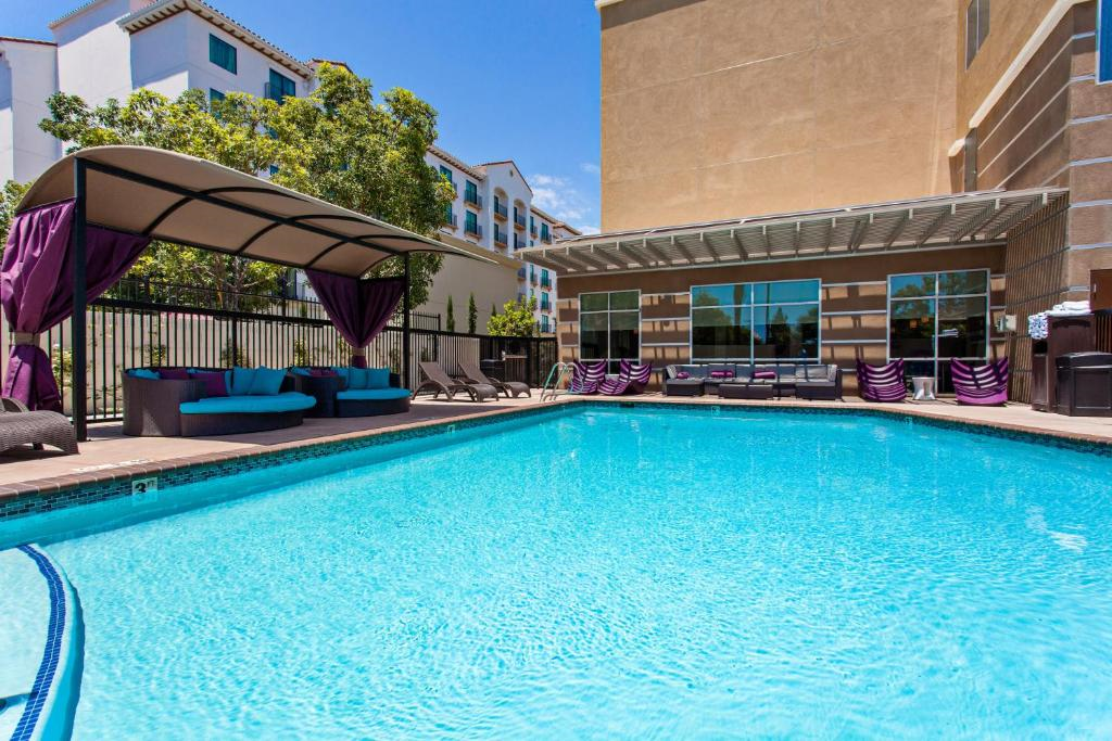 Outdoor pool area with clear blue water, surrounded by lounge chairs and a covered seating area. Building and trees in the background under a clear sky.