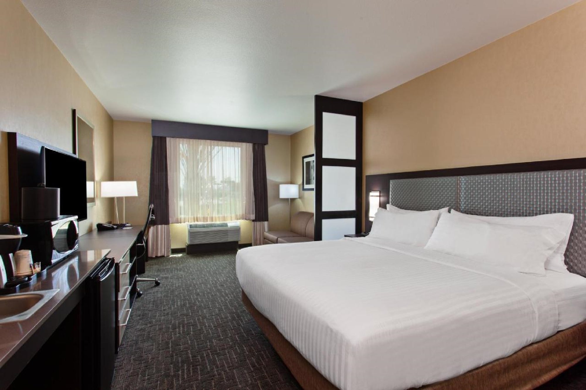 Hotel room with a king-size bed, TV, desk, armchair, and window with curtains. Neutral tones and modern furnishings.