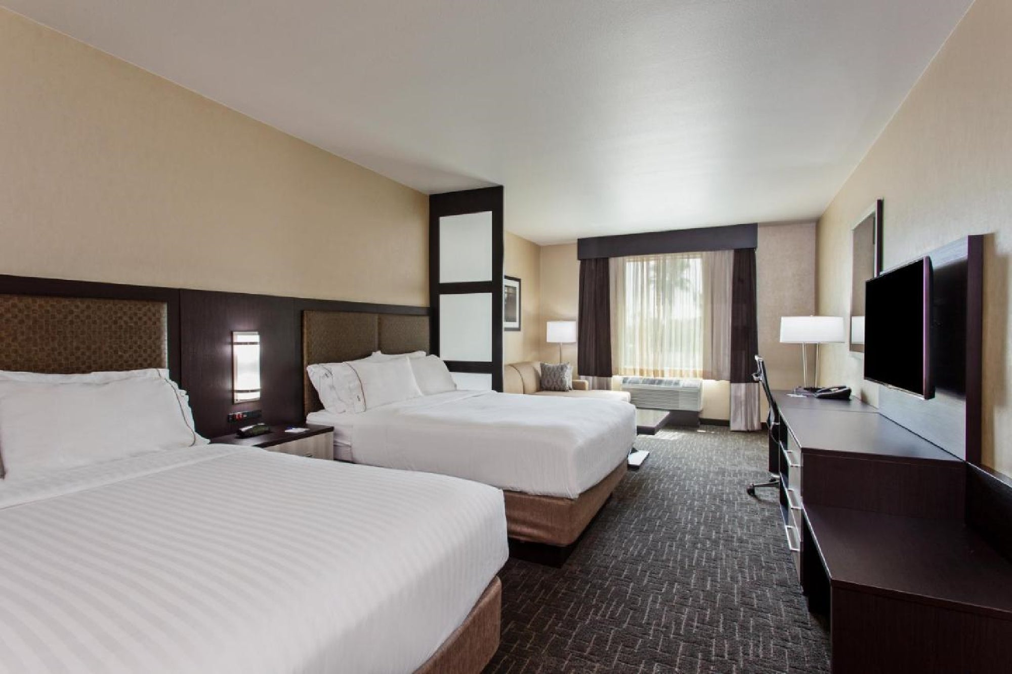 Hotel room with two large beds, a sofa by the window, a flat-screen TV, and a dark wooden desk. The room is softly lit with neutral-colored walls and carpets.