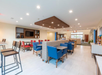 Modern communal area with tables, chairs, and a TV on the wall. The space features blue and red seating, wooden accents, and bright lighting.