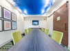 A modern conference room with a long table, green chairs, wall art, a TV, and a sliding wooden door. Blue ceiling panels add color to the room.