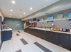 Hotel breakfast buffet area with counters, dishes, toaster, and chafing dishes. Signs on the wall feature breakfast-related messages. Walls are light green, and the floor is tiled.