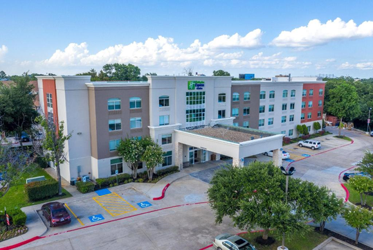 Four-story hotel building with a modern design, surrounded by a parking lot and trees under a partly cloudy sky.