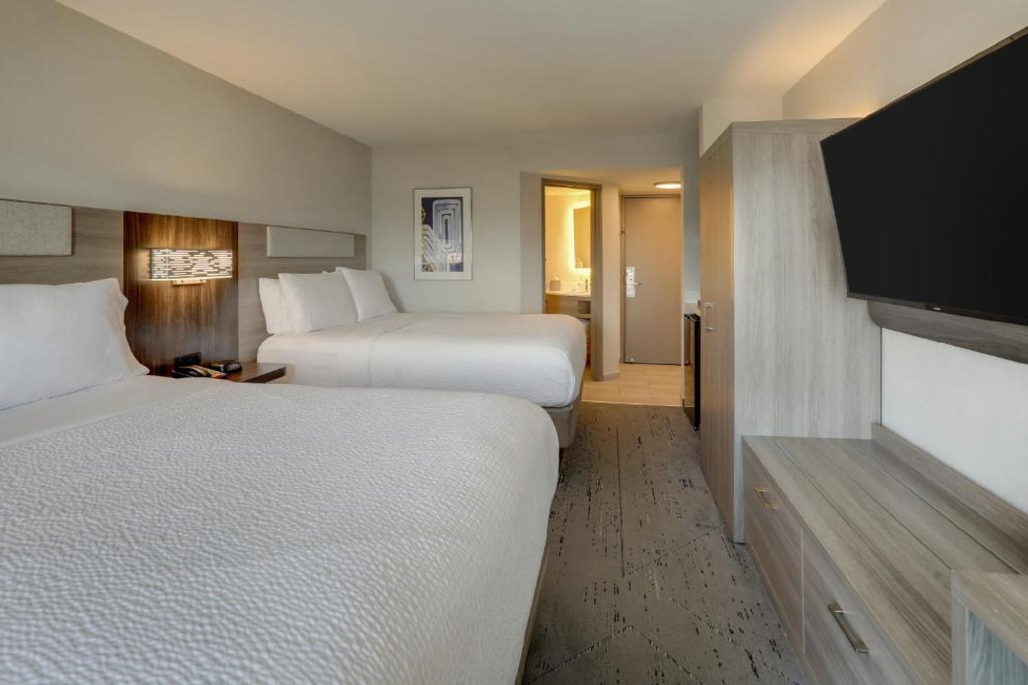 Hotel room with two large beds, wall-mounted TV, wooden furniture, and a view into the bathroom. Neutral color scheme with soft lighting.
