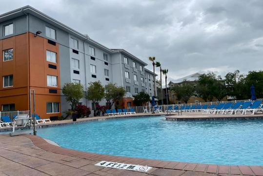Outdoor swimming pool adjacent to a multi-story hotel, surrounded by lounge chairs and trees, under a cloudy sky.