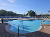 Outdoor swimming pool with clear water, surrounded by lounge chairs and umbrellas. A fountain is visible in the background under a blue sky.