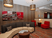 A lounge area with a large  curved padded bench, a coffee table, red accent chairs, red and gray striped carpet, and abstract art on the walls.